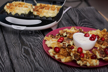 Waffles with sour cream and cranberries. Lie on a plate. Next to it is a waffle maker with waffles. On black pine boards.