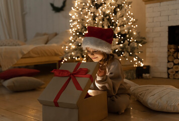 Christmas spirit. Happy little smiling girl opening gift box sitting on the floor near the...