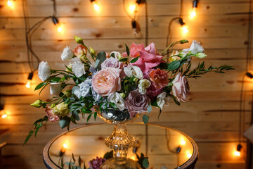 Delicate and very beautiful wedding bouquet of roses, greenery, eustoma standing on mirror table. Bridal trendy flowers in pastel colors on the background of the evening lighting bulbs