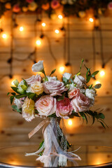 Delicate and very beautiful wedding bouquet of roses, greenery, eustoma standing on mirror table. Bridal trendy flowers in pastel colors on the background of the evening lighting bulbs