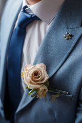 Boutonniere of beige rose flower and greenery attached to the blue suit of groom. Wedding details and floristics