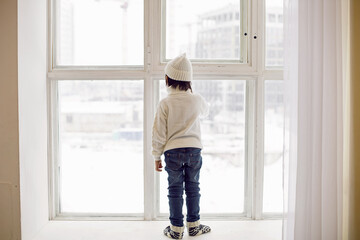 boy in a white sweater and hat stands at the window in winter in large apartment on Christmas day