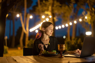 Caucasian woman watching something on laptop computer at evening time outdoors. Young woman sitting at table, eating fruits and drinking wine. Concept of weekend, rest and vacation. Idea of leisure