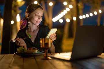 Caucasian woman watching something on laptop and phone at evening time outdoors. Young woman sitting at table, eating fruits and drinking wine. Concept of weekend, rest and vacation. Idea of leisure