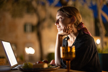 Caucasian woman watching something on laptop computer at evening time outdoors. Young woman sitting at table, eating fruits and drinking wine. Concept of weekend, rest and vacation. Idea of leisure