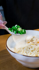 Gloved hand hold a spoon of chopped green onions over a bowl of noodles, safe catering
