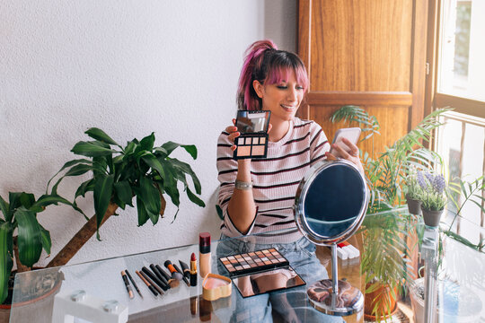 Smiling female influencer showing make-up palette while vlogging through mobile phone at home