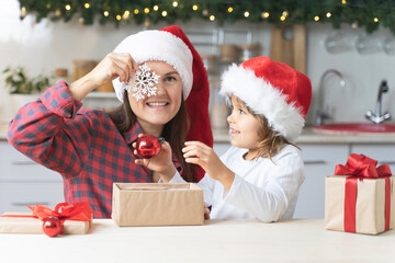Fototapeta na wymiar Happy mother and daughter in Santa hat with Christmas gifts. Cheerful family celebrates Christmas at home. Funny mom and little girl open gift boxes indoors
