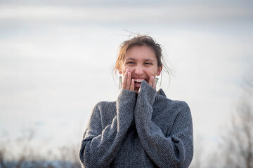 Outdoor cold winter lifestyle portrait of as single happy young woman wearing gray sweater smiling with hands on her face with white background with white_space