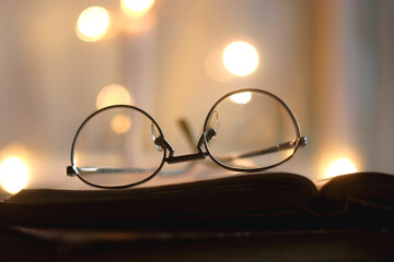 Fototapeta na wymiar Open book and reading glasses. Candles and fairy lights in the background. Selective focus.