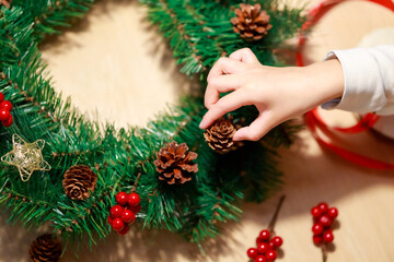 Child's right hand putting pine cone to decorate Christmas wreath on wooden background with decorative red ribbon, golden stars and red berries.