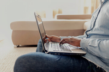 Fototapeta premium Men is typing on laptop computer keyboard. Close up of black man hands typing on a laptop sitting on the carpet at home. Shot of an unrecognizable man using a laptop at home. Technology concept.