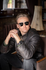 Obraz premium Sexy Blond Man in Stylish Sunglasses and a Leather Jacket Sits in an Armchair with Folded Hands. Close-up Portrait. Vintage Interior Background