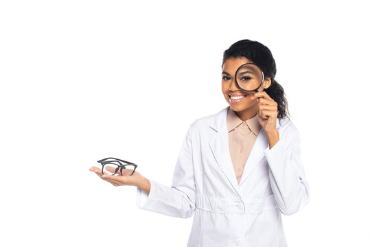 Smiling african american oculist holding eyeglasses and magnifying glass isolated on white.