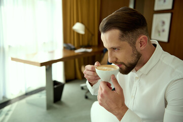 Confident attractive handsome young businessman drinks freshly brewed coffee for good start of the day, sitting in hotel room during his business trip