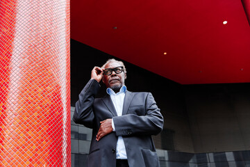 Senior male professional adjusting eyeglasses while standing by red column