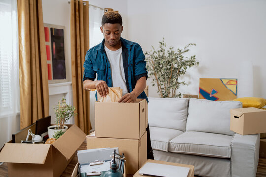 Unpacking cardboard boxes after moving, man cleans new apartment cluttered with things from previous home, looks at items, planning decorations, living room furnishings