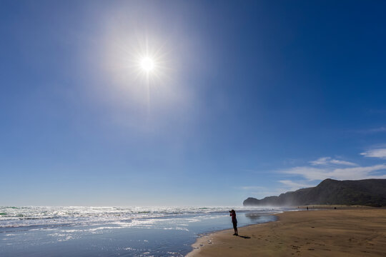 Silhouette female tourist photographing while standing at Piha Beach against clear blue sky, Auckland, New Zealand