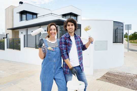 Smiling couple with paint roller standing in front of new house