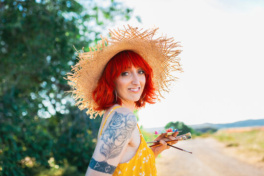 Smiling tattooed woman in straw hat holding paintbrushes and palette