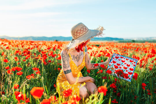 Tattooed woman holding painting amidst poppy flowers on field