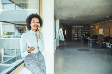 Female professional gesturing while talking on smart phone near glass window at office