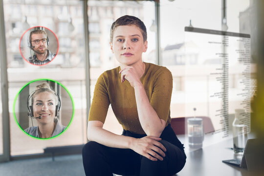 Businesswoman with hand on chin sitting on desk with male and female colleague icons in corner