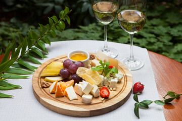 Cheese plate: sliced cheese of different kinds, honey, and fruit on a wooden tray. Next to the glasses with white wine.