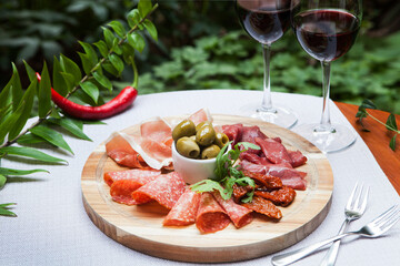Meat plate: sliced prosciutto, salami, other meats, and green olives in the center on a wooden tray. Glasses of red wine.