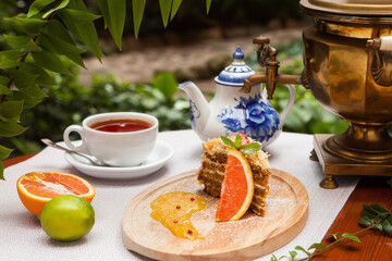 A piece of honey cake, on a wooden plate with fruit. A cup of tea and a samovar in the background.
