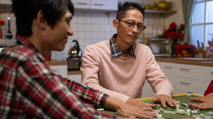 selective focus of scheming father staring at other players while mixing mahjong pieces on table. playing tile game with family at night during chinese lunar new year