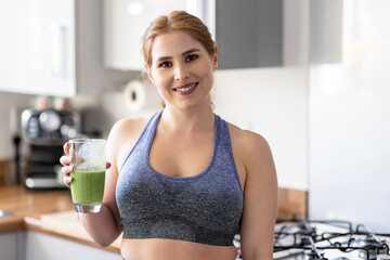 Beautiful smiling redhead woman in sports clothing holding healthy milkshake glass in kitchen