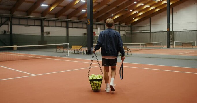 A man walks onto the tennis court, carrying a basket full of tennis balls