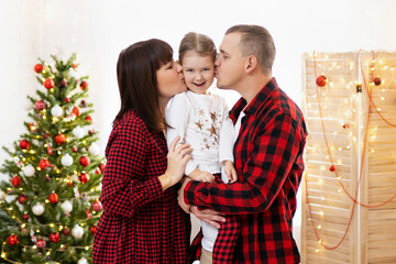 Parents holding girl on hands and kissing her near Christmas tree
