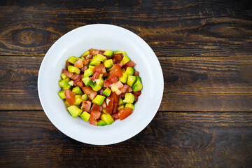 White plate with avocado and tomato salad on brown wooden surface, top view, flatlay