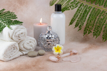 Fototapeta na wymiar Spa still life treatment composition on massage table in wellness center. Twisted hot towel with aromatic candles and face roller gua sha on beige background. Aroma therapy setting. 