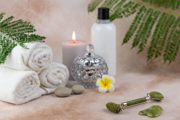 Spa still life treatment composition on massage table in wellness center. Twisted hot towel with aromatic candles and face roller gua sha on beige background. Aroma therapy setting. 