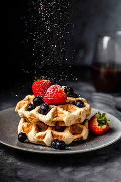 Powdered sugar falling on plate of fresh waffles with strawberries and blueberries