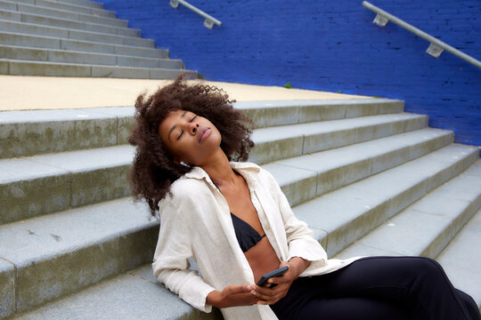 Young Afro Woman With Eyes Closed Listening Music Through In-ear Headphones Sitting On Steps