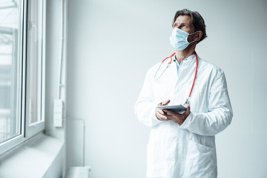 Male doctor wearing protective face mask holding digital tablet while standing by window in clinic