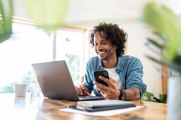 Smiling male professional holding smart phone while using laptop on table at home
