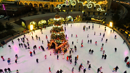 Top view of people skate on ice rink around christmas tree in park on winter holidays. Active...