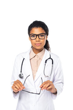 African american ophthalmologist in white coat holding eyeglasses and looking at camera isolated on white.