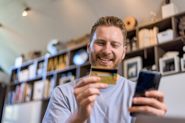 Low angle shop of handsome young adult smiling man holding credit card and using phone doing online...