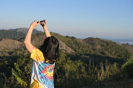 A female tourist wearing a plaid long-sleeved shirt and a yellow bag uses a camera to record the misty morning atmosphere. Covering the sky,  trees in the soft sunlight while traveling in the mountain