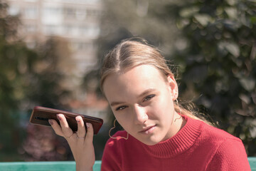 young beautiful girl 16 years old, in a red sweater, sits in the park with a phone and listens to...