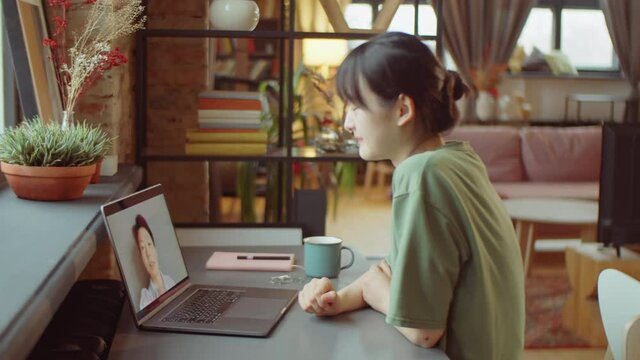 Side view of young Asian woman sitting at desk at home and talking friend on online video call on laptop