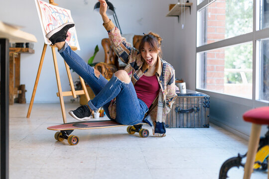 Cheerful young woman sitting on skateboard in artist's studio