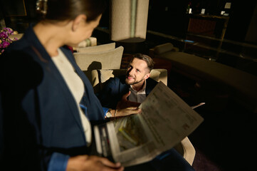 Focus on a handsome Caucasian businessman looking at his blurred female business partner, sitting in front of him and reading newspaper in a hotel lobby