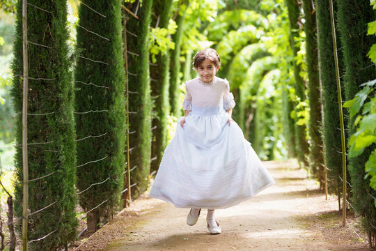 Cute girl in white communion dress running on footpath in green garden